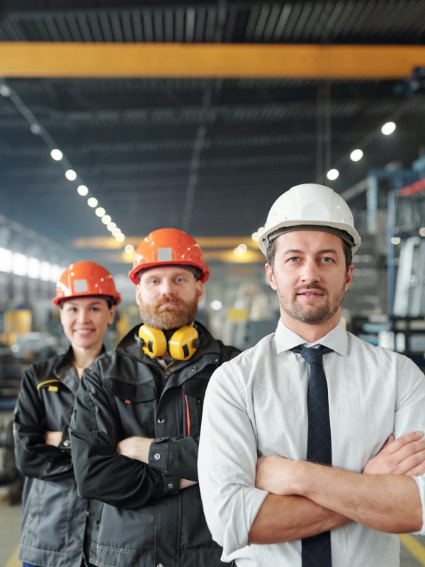 Young cross-armed leader of engineering team and his successful trainees in workwear standing in front of camera in factory